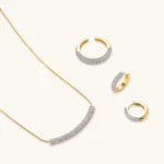 Gold-plated curved necklace, hoop earrings, and adjustable ring with pavé cubic zirconia stones on cream background.