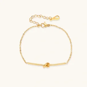 Delicate gold chain bracelet with minimalist bar and centered knot detail on cream background