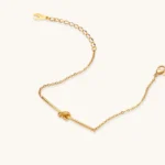 Delicate gold chain bracelet with minimalist bar and centered knot detail on cream background