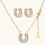 Gold-plated horseshoe necklace and earring set with baguette and round crystals arranged in geometric pattern on white background.